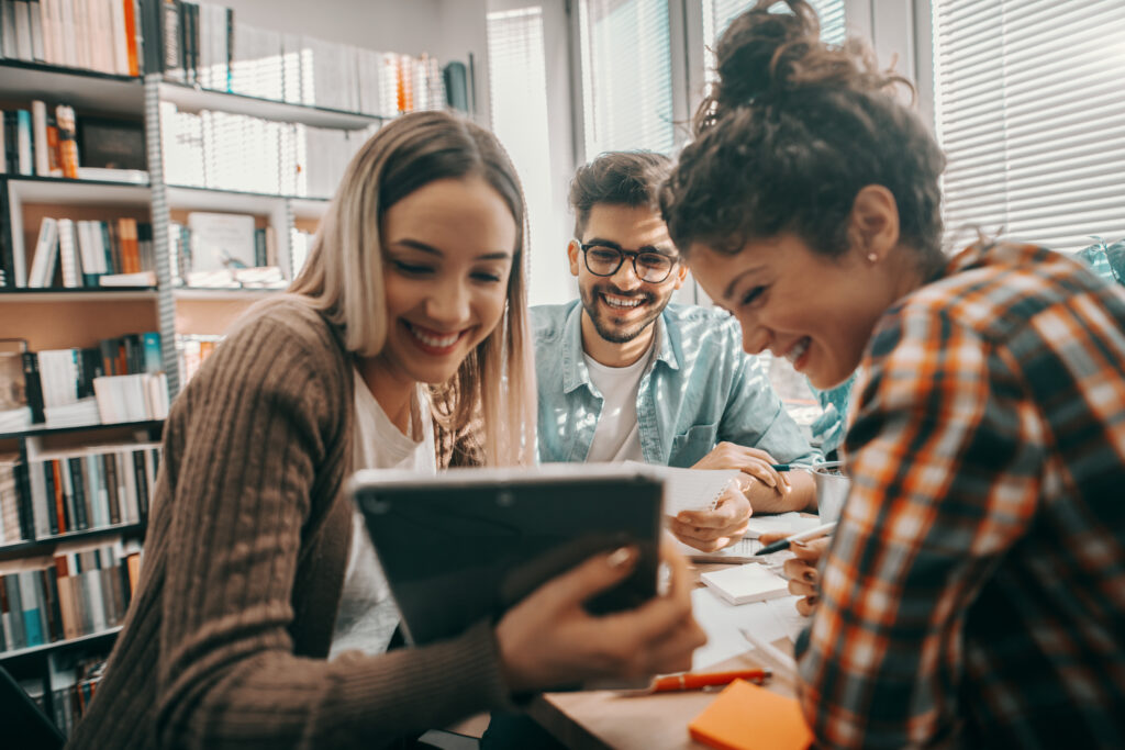 Three happy students dressed casual using tablet for school project and sitting at desk in library. Contrato digital na educação: quais as vantagens?