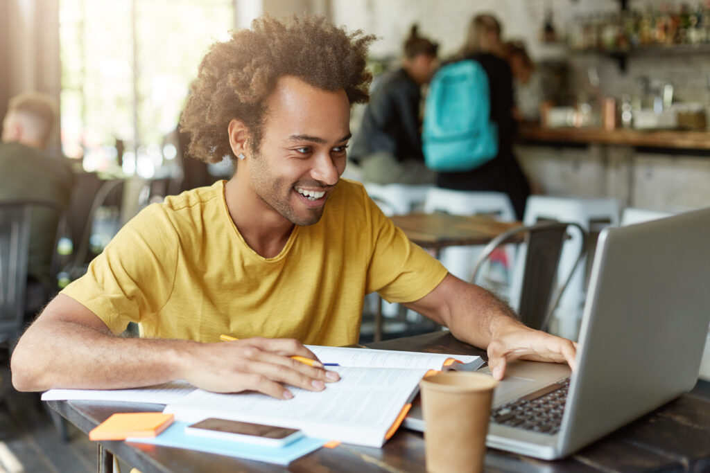 Indoor shot of happy student male with curly hair dressed casually sitting in cafeteria working with modern technologies while studying looking with smile in notebook receiving message from friend Diploma digital, ensino à distância e outras tendências da Educação para 2022