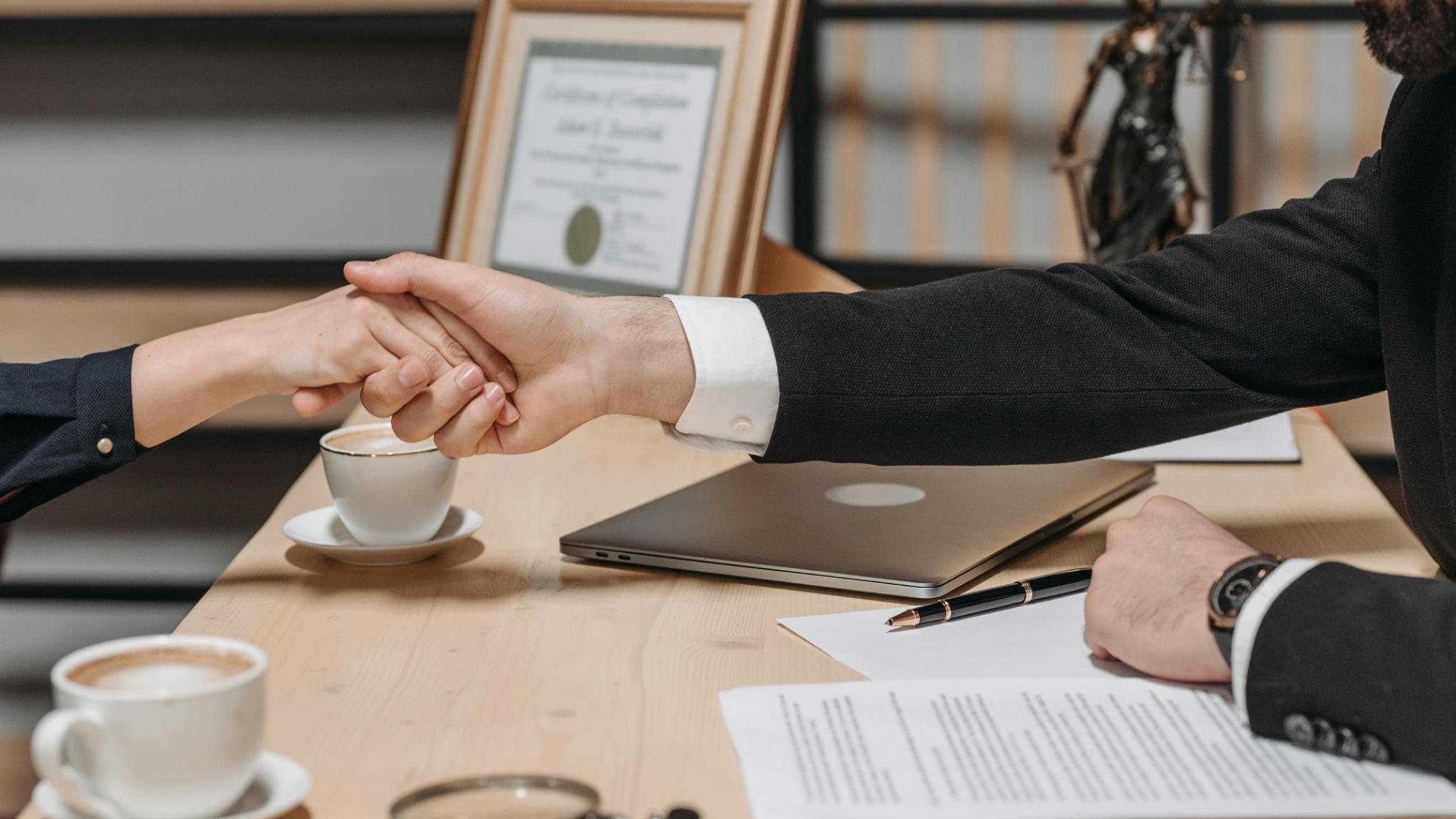 handshake-law-landscape Pessoa aperta a mão de outra pessoa em um escritório de advocacia com documentos, laptop e estátua da Justiça ao fundo.